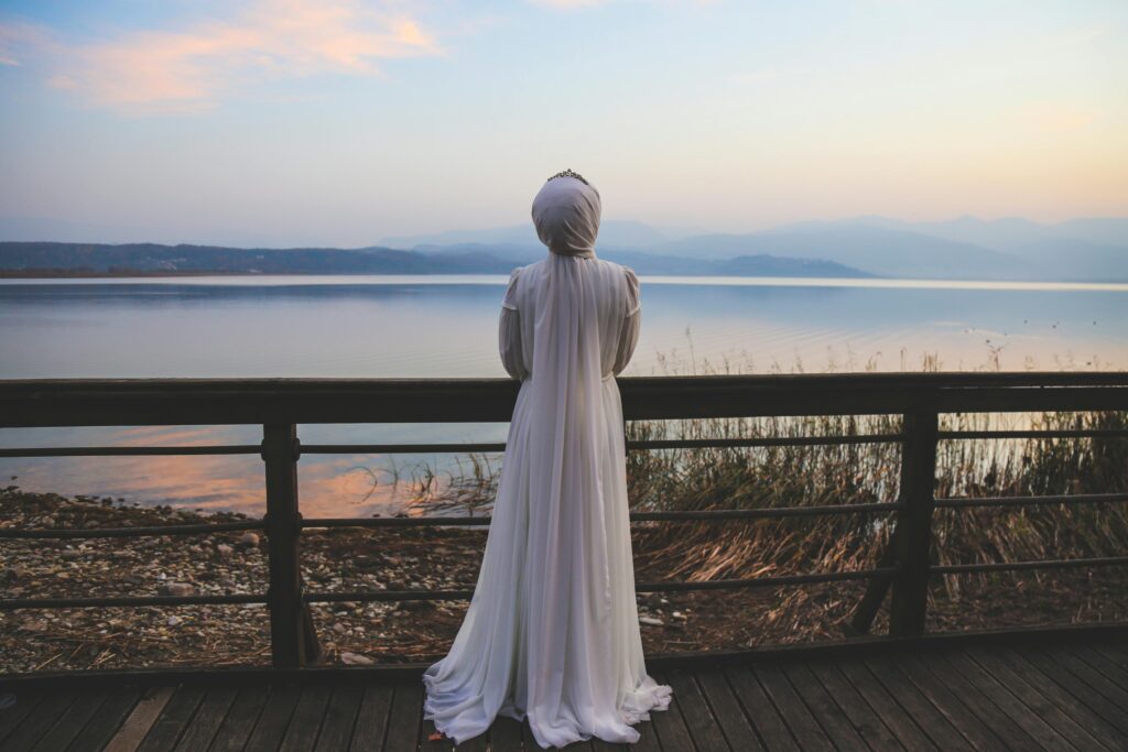 pexels-photo-11034913-11034913 A bride in a white dress gazes over a peaceful lake at dawn, reflecting tranquility.