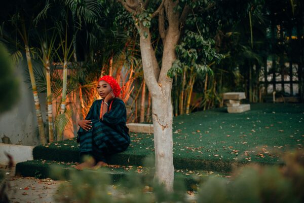 A woman in traditional attire sitting amidst lush greenery and a tree in a garden area.