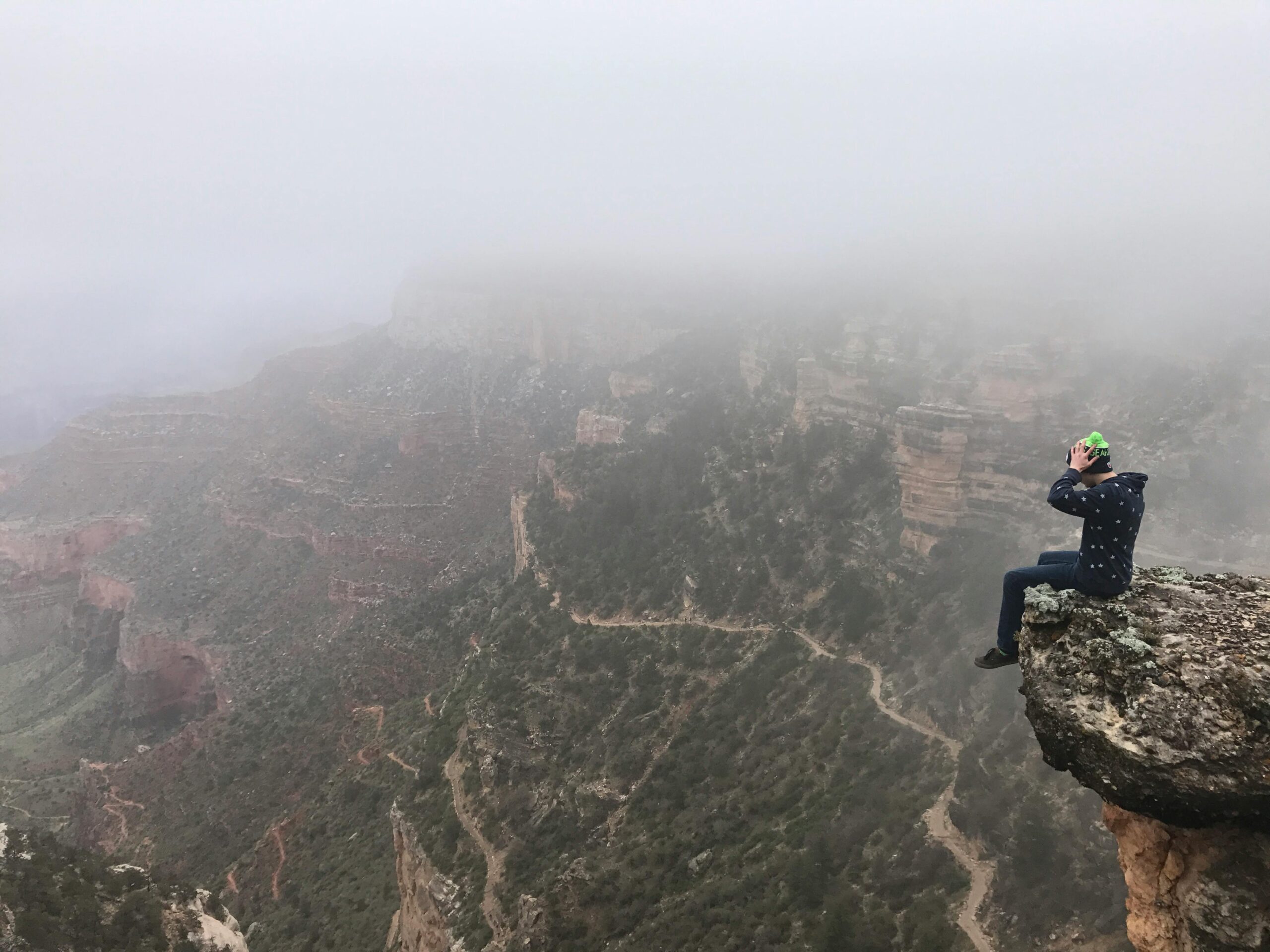 A daring hiker sits on a foggy cliff edge of the Grand Canyon, capturing the serene magnitude of nature.