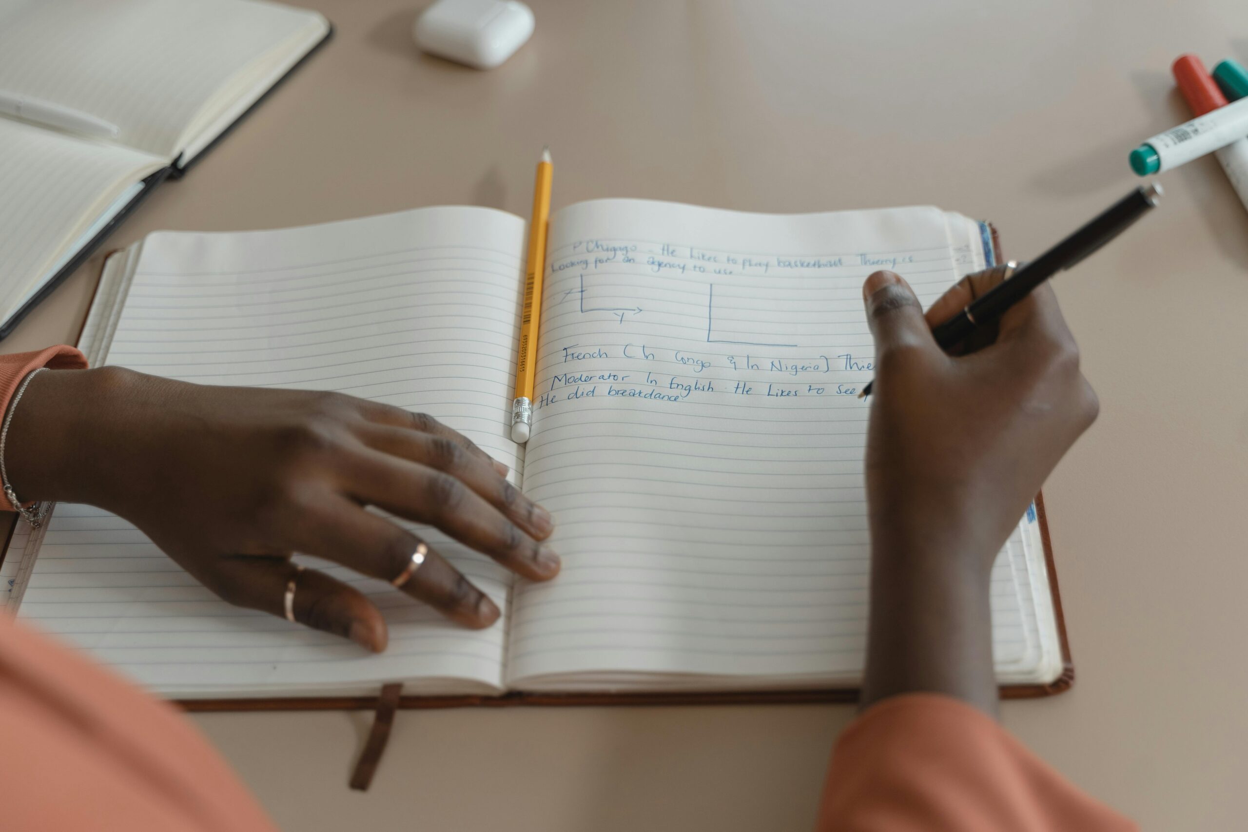 Focused close-up of hands writing in a notebook with a pen and pencil on a table.