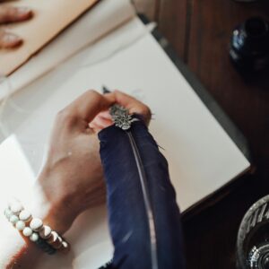 High angle of crop anonymous female author with feather and blank notebook at desk in sun ray