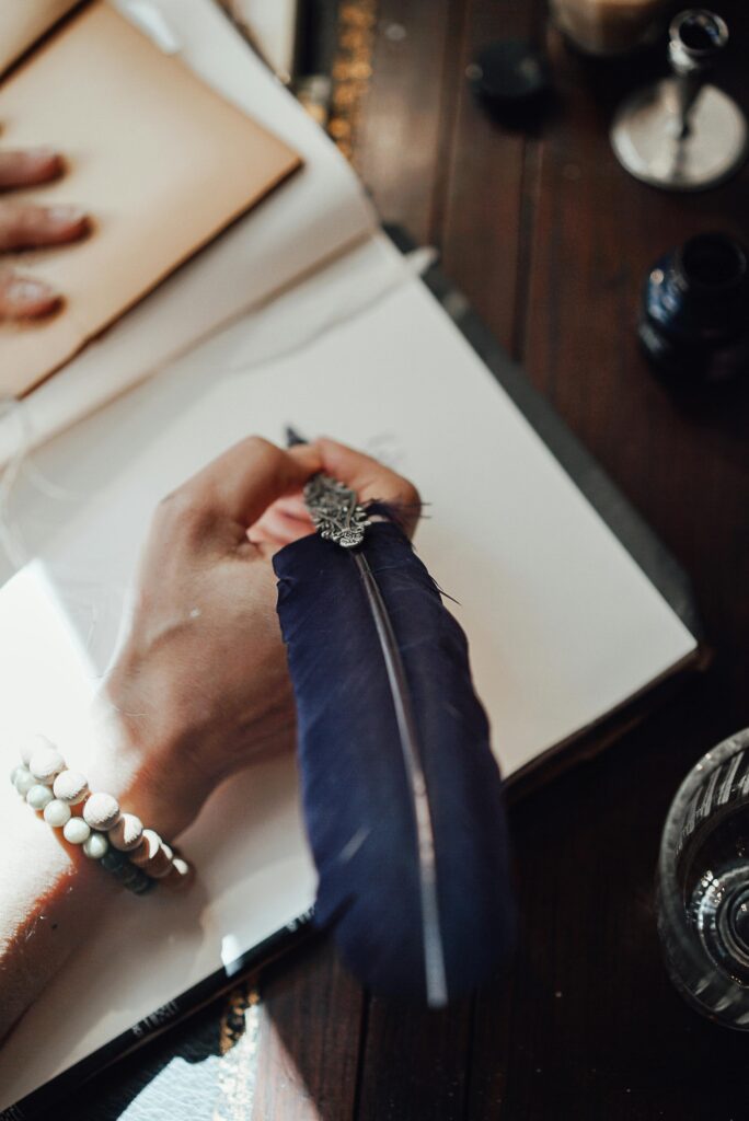 pexels-photo-7080688-7080688 High angle of crop anonymous female author with feather and blank notebook at desk in sun ray